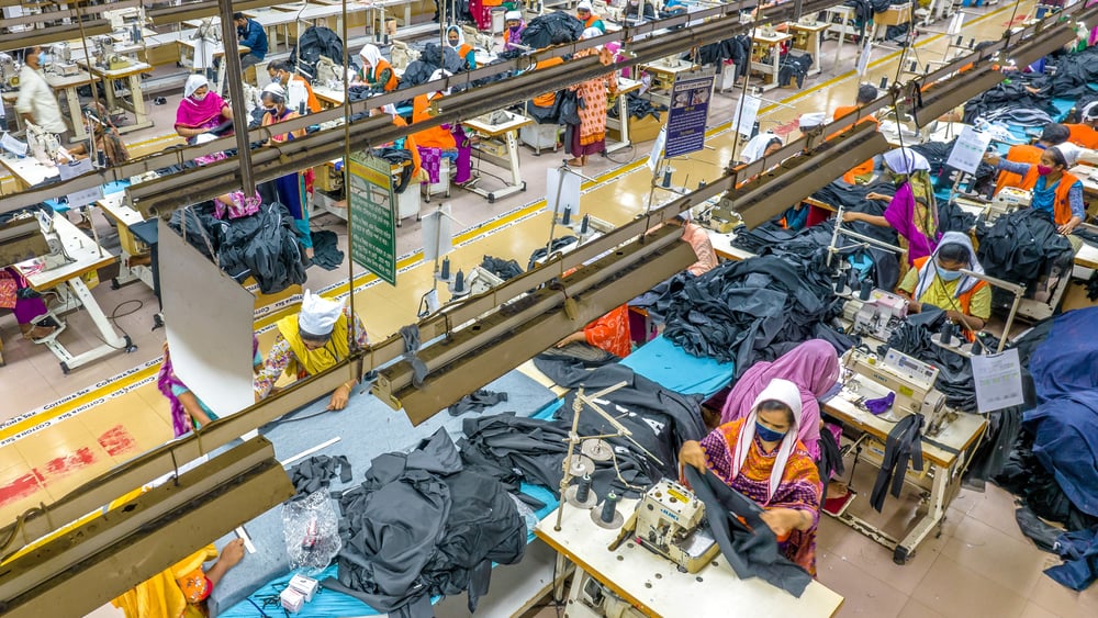 Näherinnen in einer riesigen Fabrikhalle vor ihren Nähmaschinen und Stoffbergen.