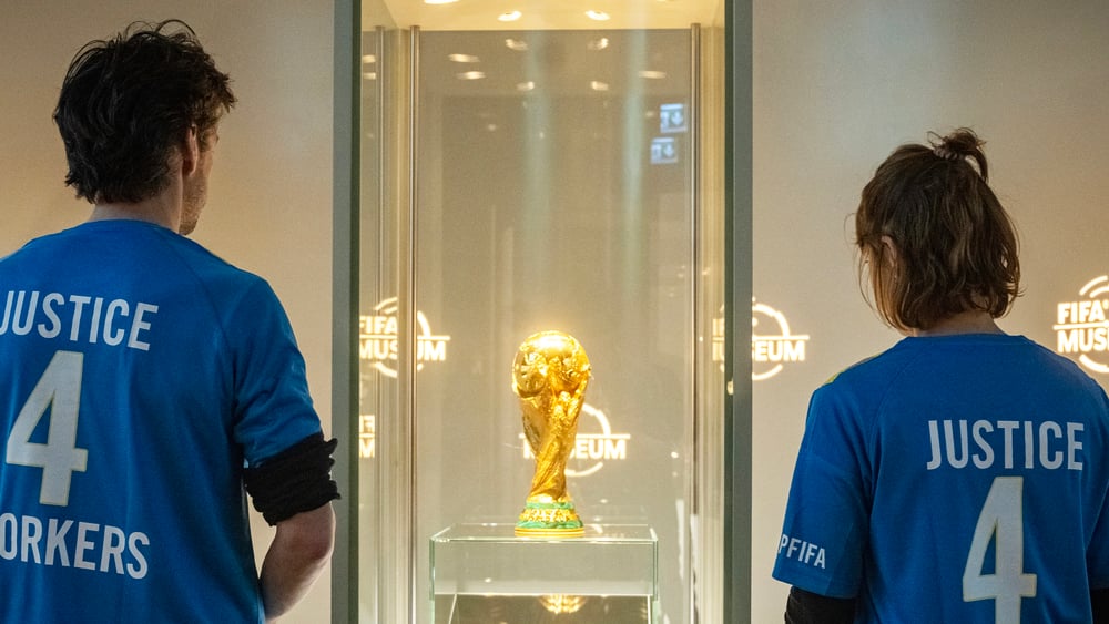 Ein junger Mann und eine junge Frau stehen im Fifa-Museum vor dem WM-Pokal mit T-Shirts auf denen Justice for Workers steht.