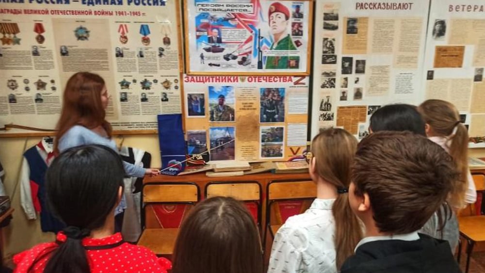 Eine Lehrerin vor einer Tafel mit Bildern und Dokumenten in Russisch, eine Zahl Kinder schaut ihr zu.