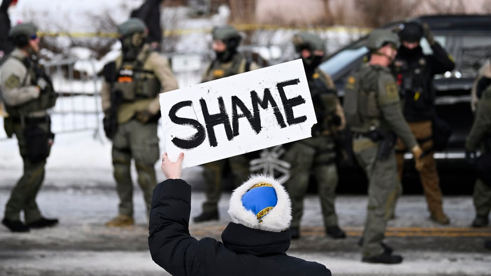 Eine demonstrierende Person mit einem Schild mit der Aufschrift «Schande» beobachtet am 7. Januar 2026 in Minneapolis, Minnesota, Einsatzkräfte, die den Tatort sichern, an dem eine Frau von einem ICE-Beamten erschossen wurde. © Stephen Maturen/Getty Images