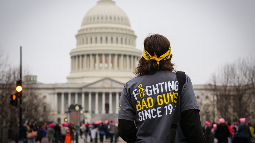 Person mit gelbem Bandana und grauem T-Shirt mit der Aufschrift ‚Fighting Bad Guys Since 1961‘ steht vor dem US-Kapitol in Washington, D.C.; im Hintergrund sind Menschen und das Kapitolgebäude sichtbar.