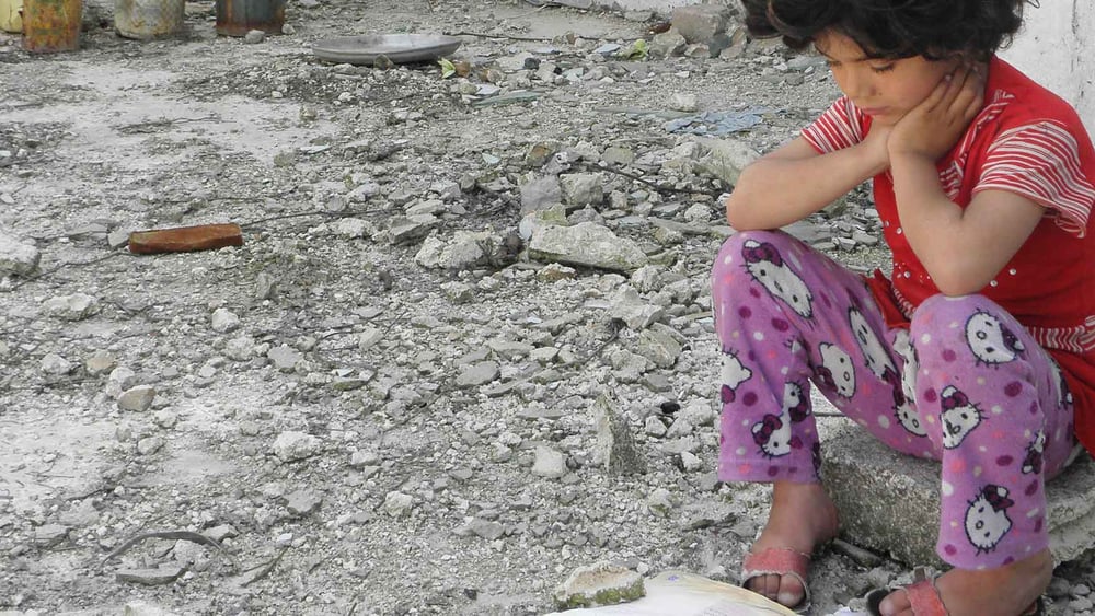 Ein syrisches Mädchen liest vor ihrem beschädigten Zuhause in Homs in einem Buch. April 2012. © Reuters/Stringer