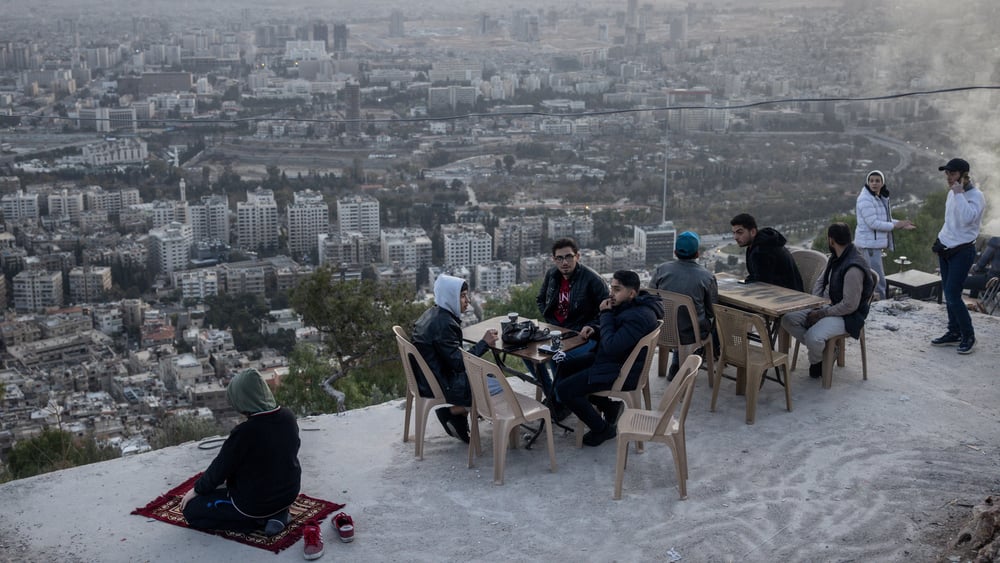 Junge Syrier*innen sitzen auf dem Aussichtspunkt an Tischen. Einer kniet auf einem Teppich und scheint zu beten.