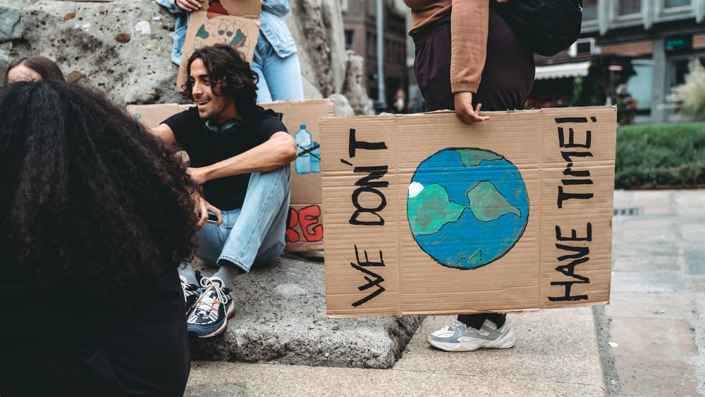 Eine Gruppe von Personen, welche am Strassenrand sitzen und Diskutieren. Zu sehen ist ein Plakat auf welcher die Erde gemalt ist. Darüber und darunter steht: We don't have Time!