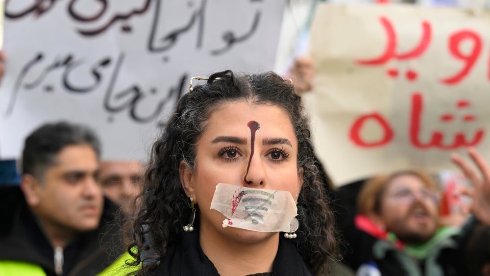 Eine junge Frau mit einem roten Punkt auf der Stirn, der einen Kopfschuss symbolisiert, und einem Pflaster über dem Mund.