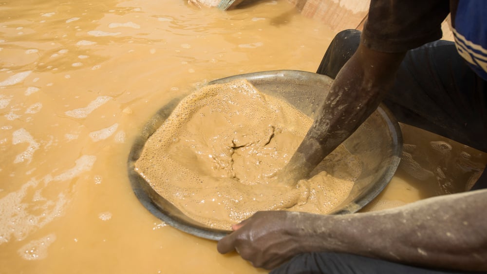 zwei Hände sind mit einer Schüssel beim Goldwaschen in braungelbem Wasser.