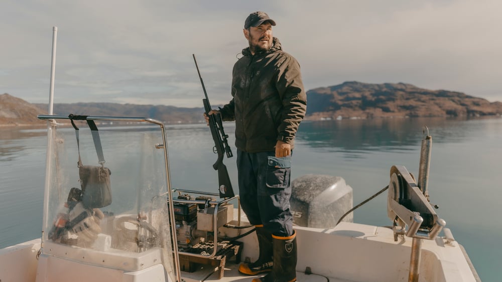 Janussen steht in Fischerkleidung auf seinem Boot mit einem Gewehr in der Hand.