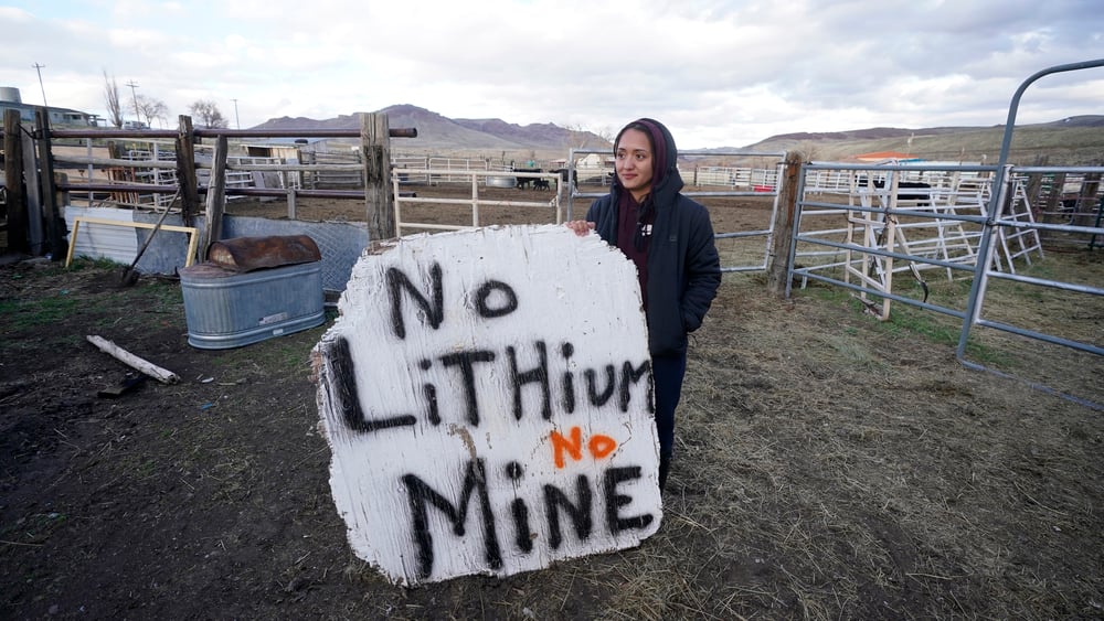 Eine junge Frau steht mit einem grossen Transparent, auf dem "No LIthium, no MIne" steht, vor etwas, das wie eine Pferdekoppel aussieht.
