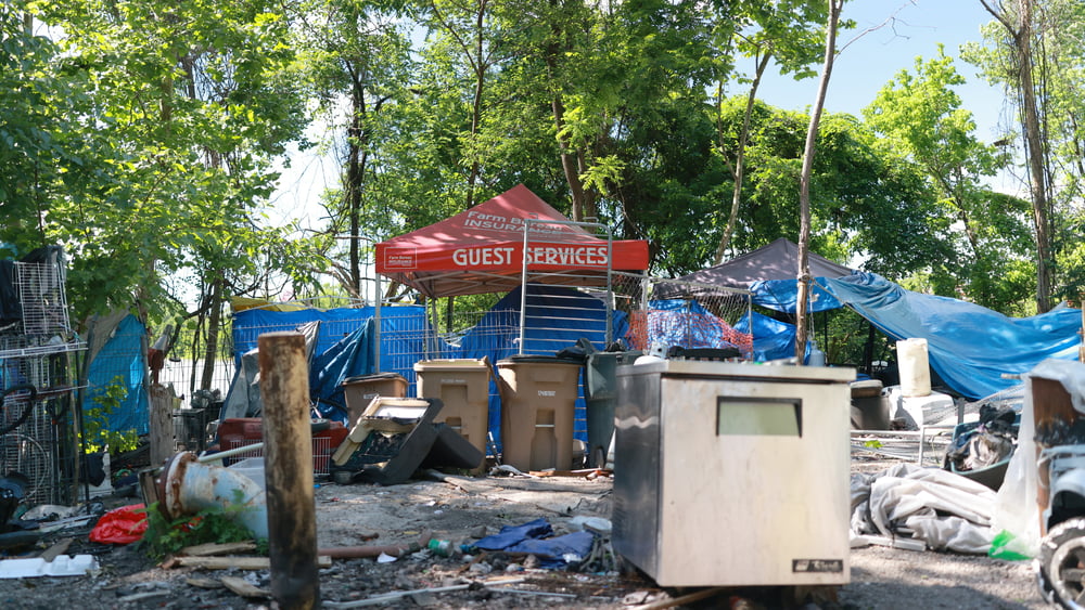 Eine Ansammlung von Planen, Abfallkübel, ein Container und ein Sonnenschirm zeigen ein Durcheinander von Gerümpel und Hüttenutensilien.