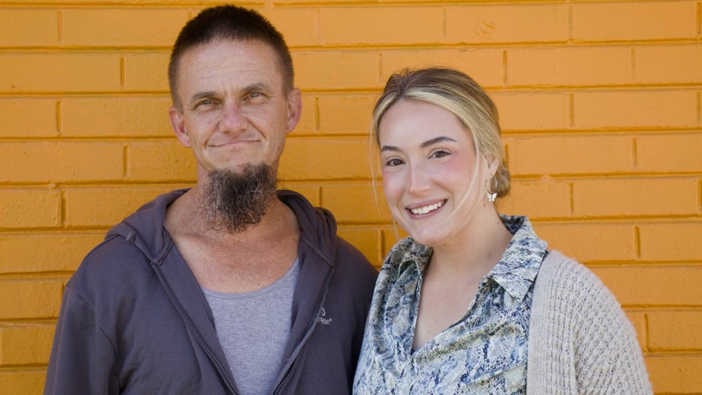 Andy und Melissa stehen nebeneinander vor einer orangen Ziegelmauer.