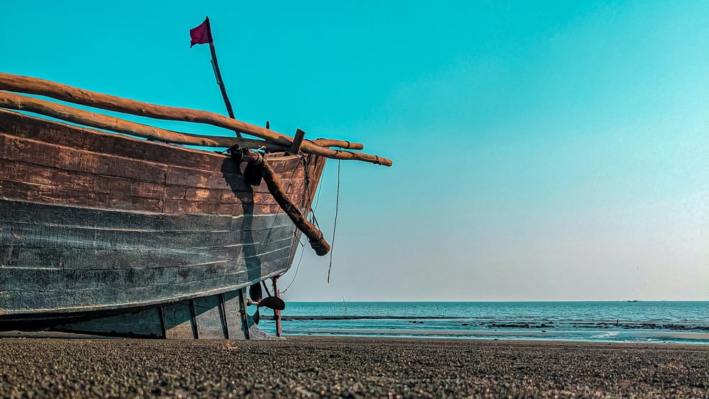 Ein altes, halb kaputtes Boot am Strand vor blauem Himmel.