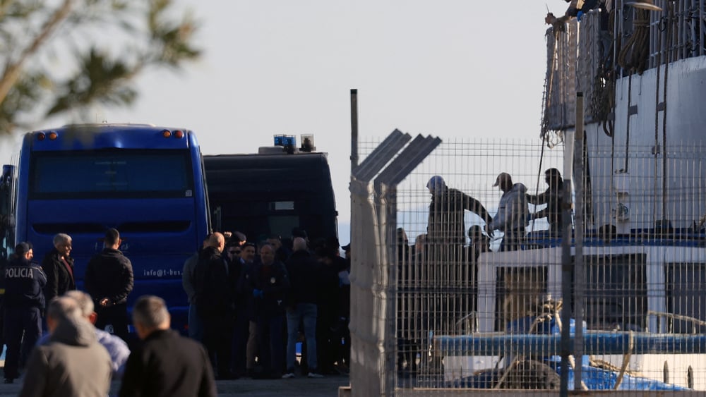 Drei Männer, einer davon offenbar in Handschellen, werden vom Schiff zu einem Wagen geführt.