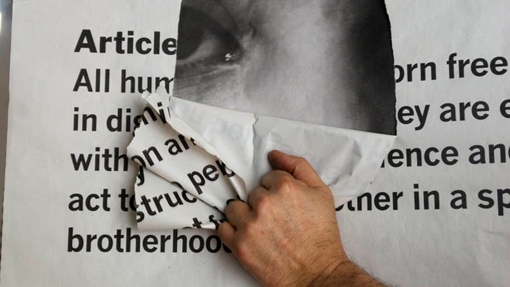 Schwarz-weiss-Bild eines Fotopanels mit dem ersten Artikel der AEMR, welches von einer Hand aufgerissen wird. Dahinter im Loch sieht man ein Gesicht.