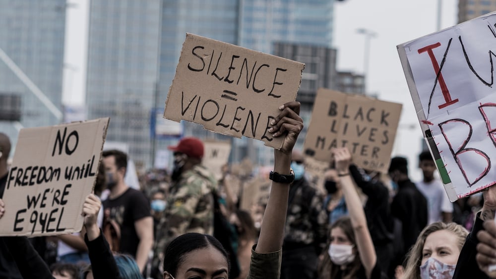 Demonstration gegen Rassismus mit aufgestreckten Armen dabei ein Transparent mit der Aussage Silence is Violence