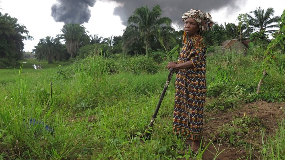 Die Bäuerin Taagaalo Christina Dimkpa Nkoo vor ihrem Land, das durch eine beschädigte Ölpipeline von Shell verschmutzt worden sei. Im Hintergrund Rauch aus der Pipeline in Kegbaara Dere, Rivers State, Nigeria. © Amnesty International