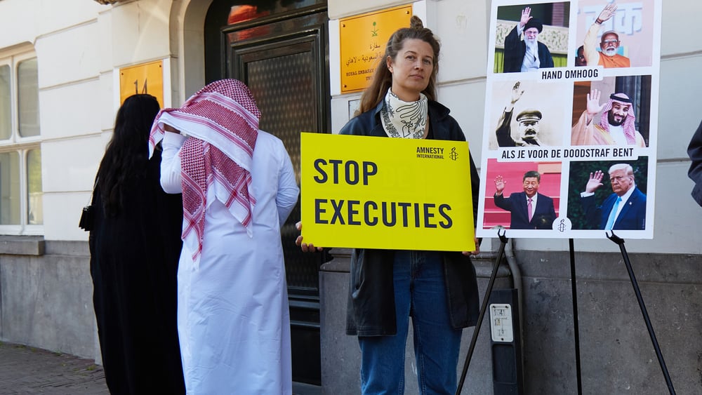 Une femme se tient devant l'ambassade, brandissant une pancarte d'Amnesty sur laquelle on peut lire en néerlandais « Arrêtez les exécutions » (stop executies)