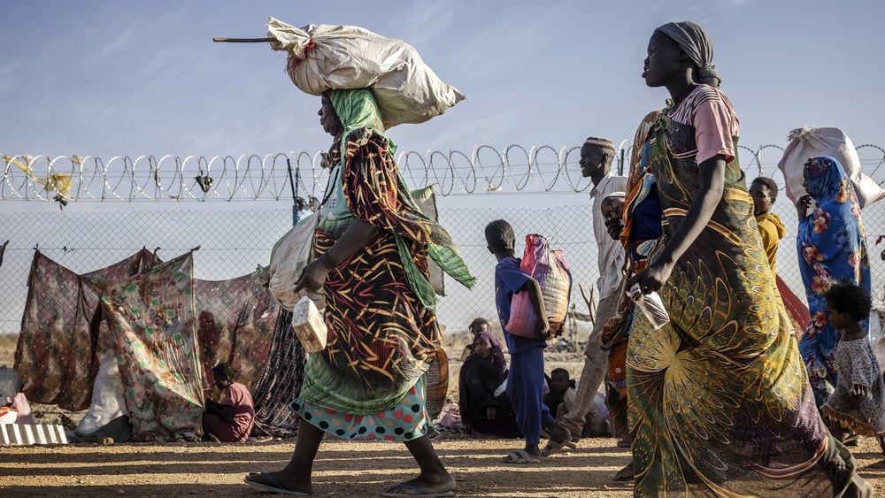 Plusieurs personnes, dont des femmes et des enfants, passent devant un terrain clôturé de barbelés. Elles portent des vêtements colorés et transportent de grands sacs et des paquets sur la tête ou dans les mains. À l'arrière-plan, d'autres personnes sont assises par terre à côté d'abris de fortune faits de tissu et de bâches, sous un ciel dégagé.