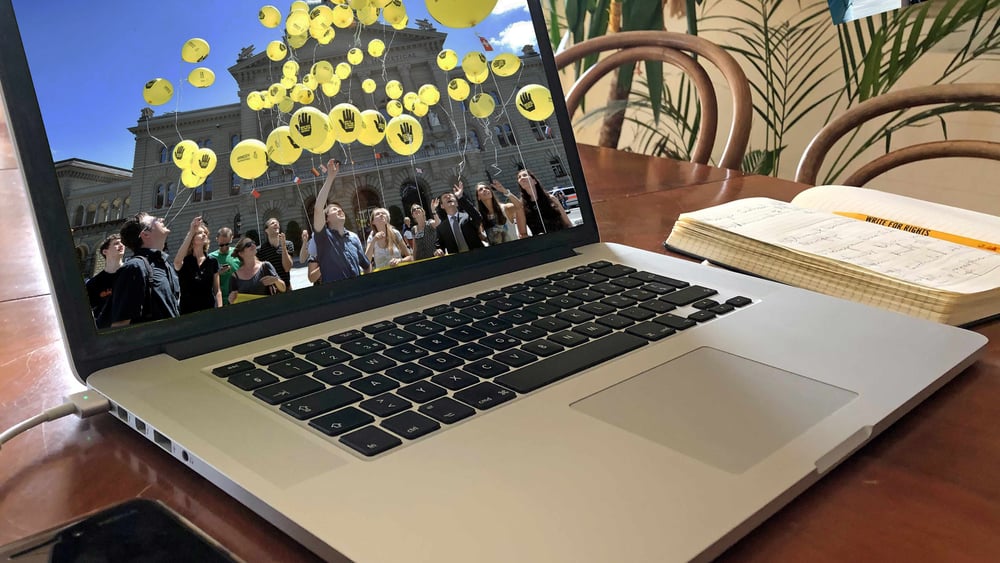 Une main tape sur un ordinateur portable dont l'écran affiche le Palais fédéral et des ballons d'Amnesty