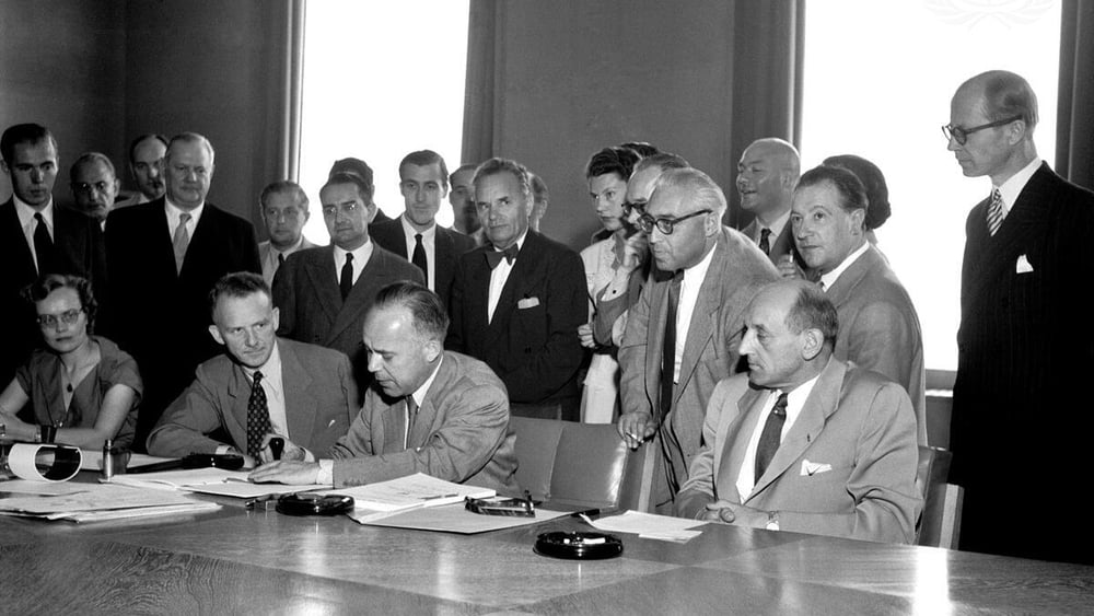 Photo en noir et blanc : plusieurs personnes sont assises autour d'une table, l'une d'entre elles signe un document.