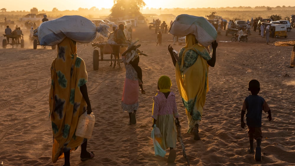 Silhouettes de femmes et d'enfants réfugiés, vus de dos, qui, au coucher du soleil, semblent suivre une caravane de personnes, de charrettes en bois et de voitures.