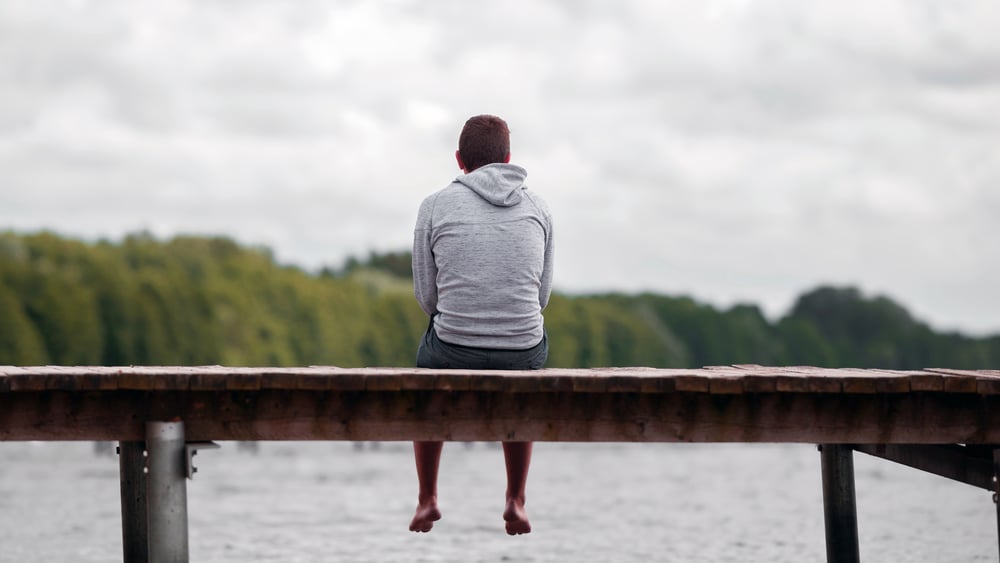 Un jeune homme en sweat à capuche et short est assis sur un ponton et regarde au loin , vu de dos.