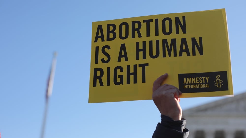 Une main tient une banderole jaune d'Amnesty International sur laquelle on peut lire « Abortion is a Human Right » (L'avortement est un droit humain).