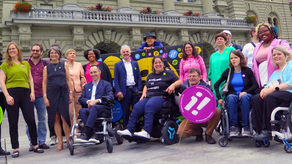 Plusieurs militant·e·x·s, dont certain·e·x en fauteuil roulant, se tiennent derrière une grande banderole sur laquelle on peut lire « Des droits humains pour tous ».