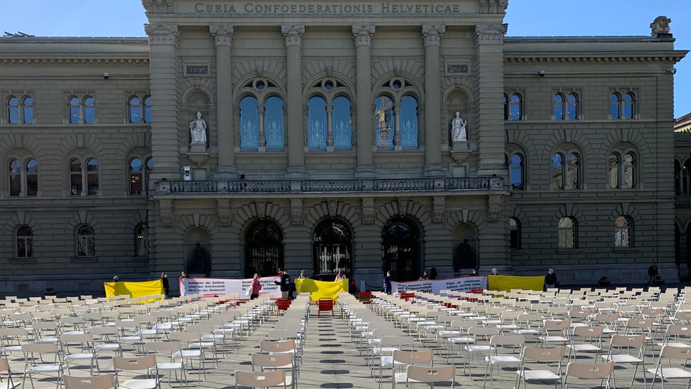 Facciata del Palazzo federale a Berna con file di sedie vuote e striscioni di protesta in primo piano.
