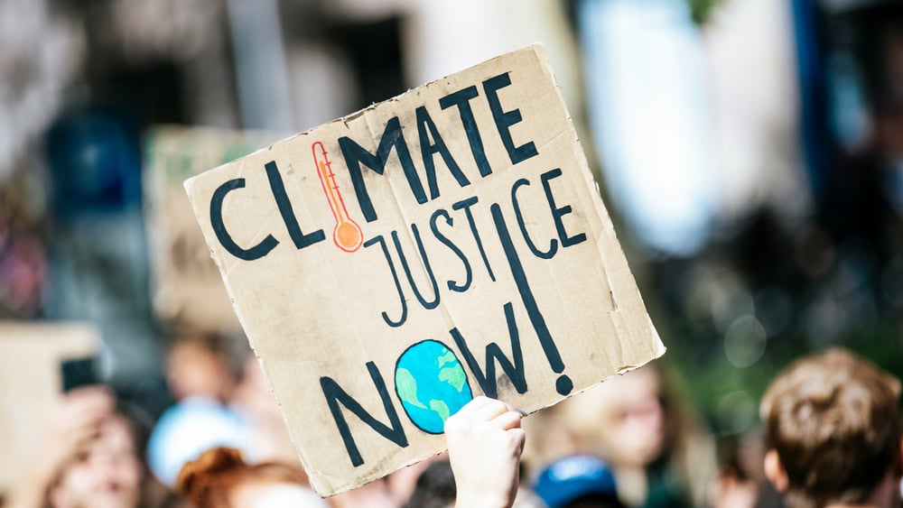 Durante una manifestazione una mano tiene un cartello con la scritta "Climate justice now!" Giustizia climatica ora!).