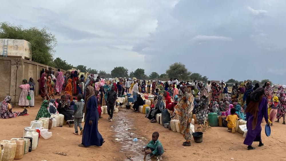 Un gran numero di donne e bambini attendono con taniche e pentole su un terreno sabbioso.