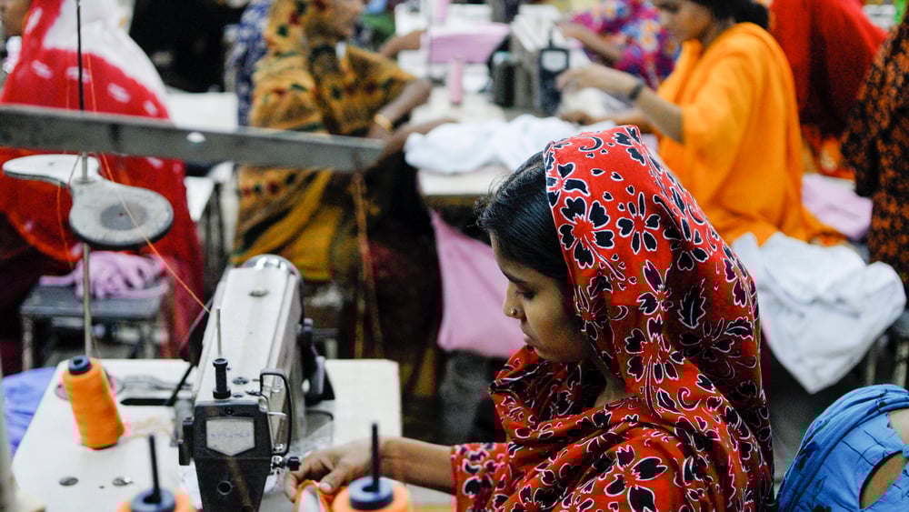 Una giovane sarta alla macchina da cucire; alle sue spalle si vedono altre lavoratrici.