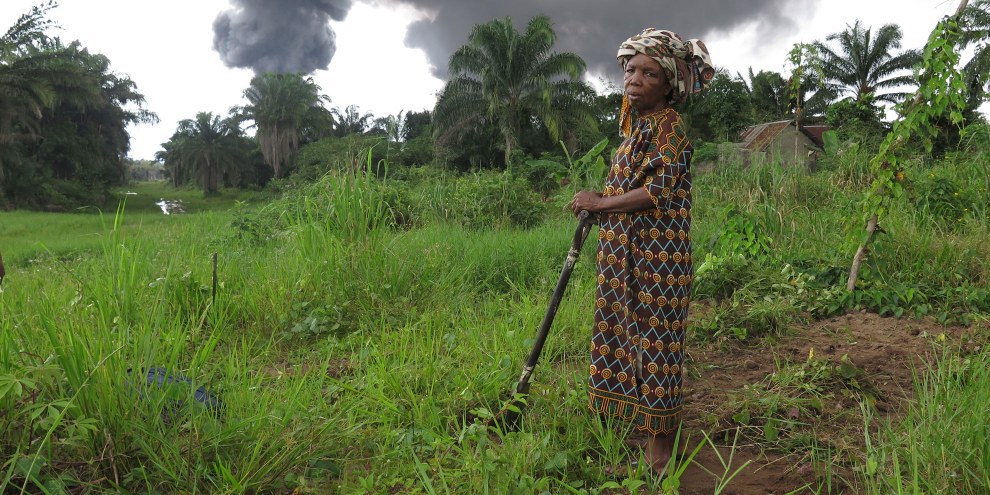 Die Bäuerin Taagaalo Christina Dimkpa Nkoo vor ihrem Land, das durch eine beschädigte Ölpipeline von Shell verschmutzt worden sei. Im Hintergrund Rauch aus der Pipeline in Kegbaara Dere, Rivers State, Nigeria. © Amnesty International